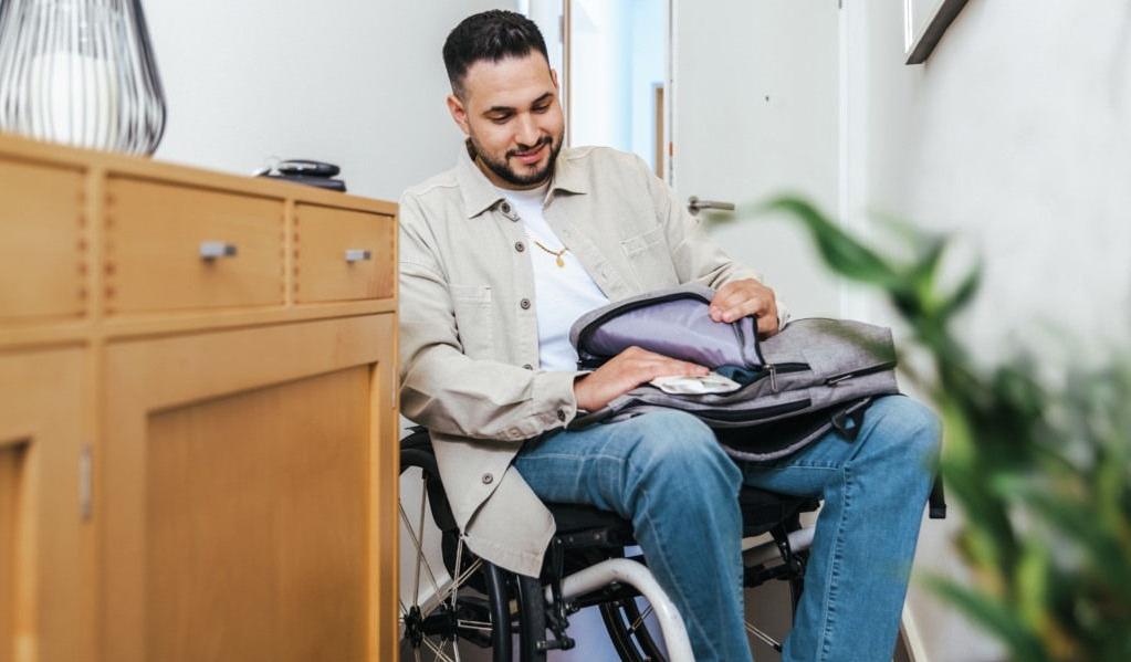 A man in a wheelchair, a bathroom in the background, holding a cathether in his hand